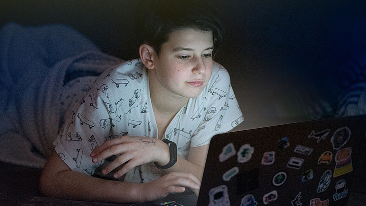 Jugendlicher liegt in einem dunklen Raum und schaut auf einen geöffneten Laptop